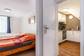 Ein Schlafzimmer mit zwei Betten, einem fenster mit Vorhängen und Blick in eine Küche mit Herd und Schränken.
