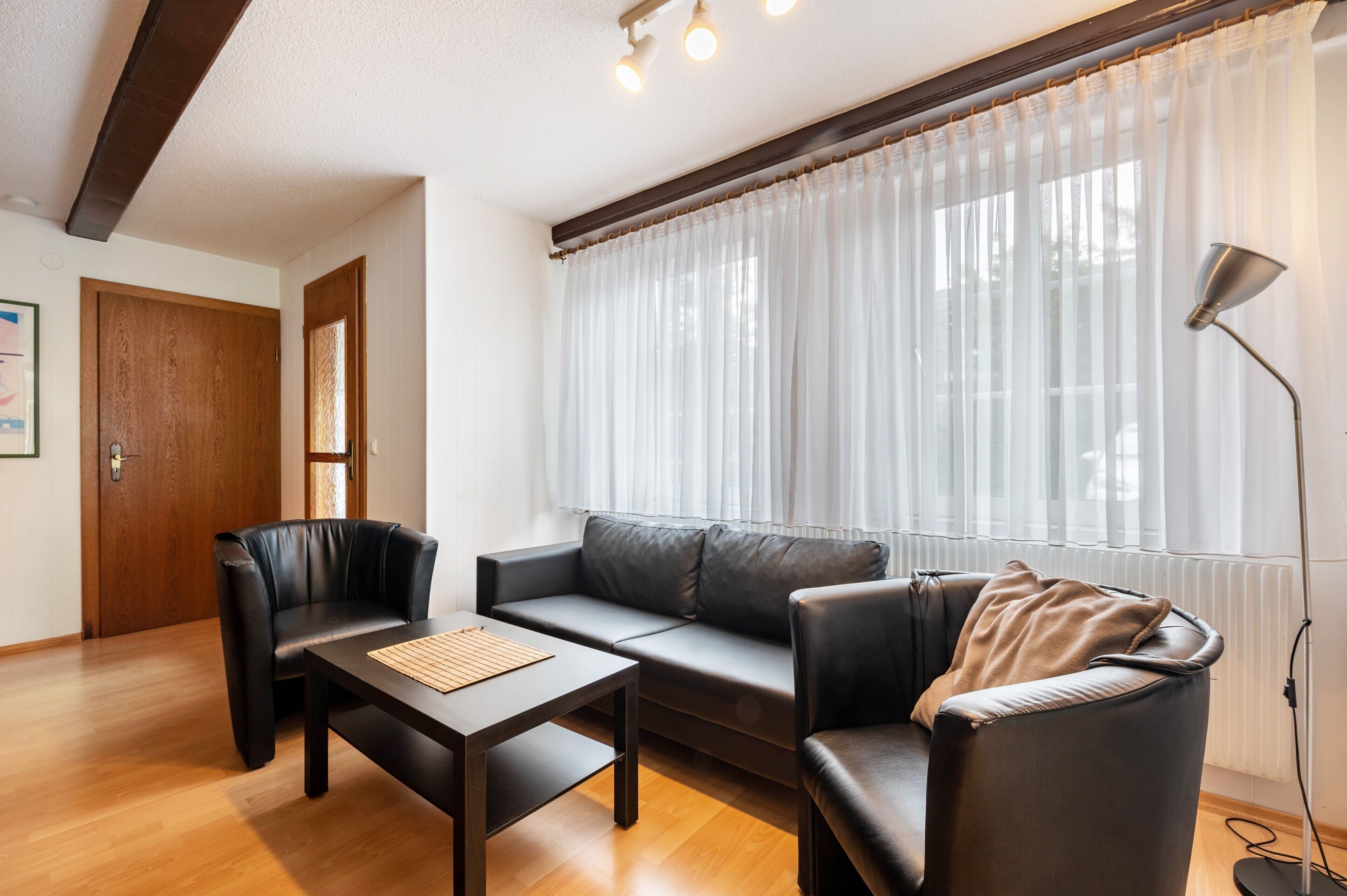 A room with a sofa, two armchairs, a coffee table, a lamp, and a window with sheer curtains.