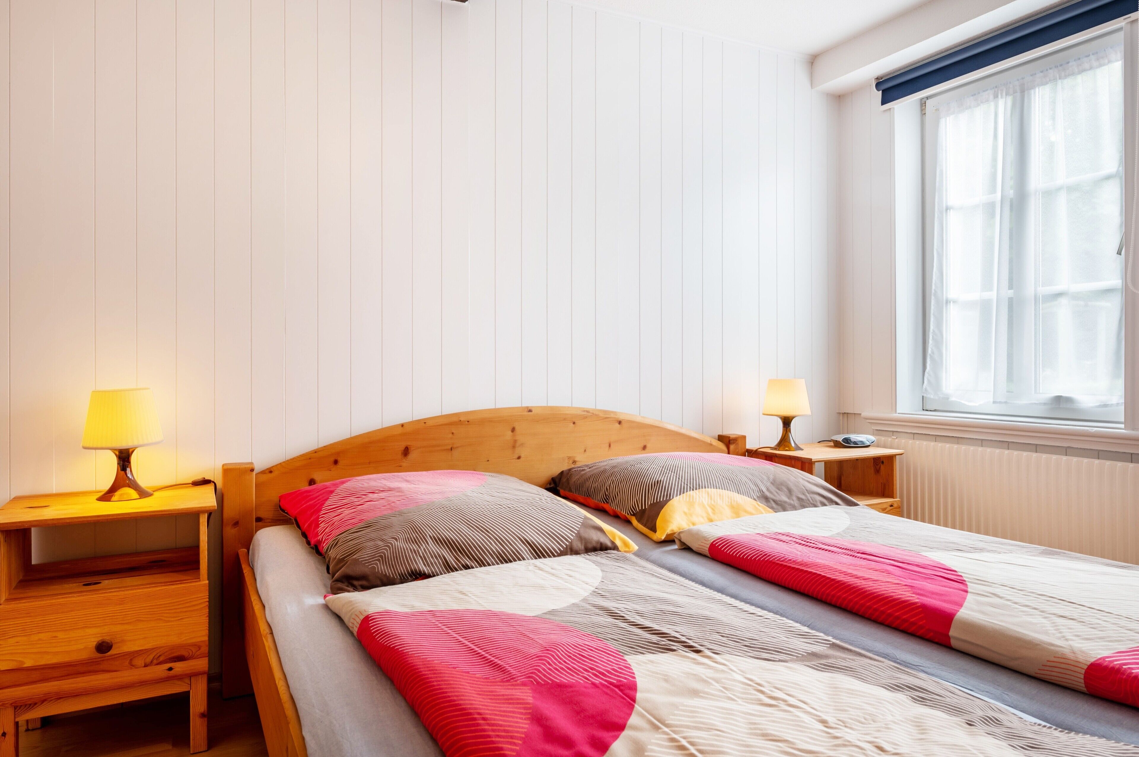 A double bed with a wooden headboard, two bedside tables with lamps, a window with curtains, and a radiator.