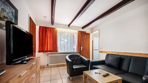 A modern living room with a flat-screen TV, a wooden cabinet, a black leather sofa, a wooden coffee table, and a window with red curtains.