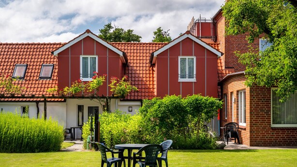 A two-story house with a red-tiled roof, a garden with a table and chairs, and a pathway leading to the entrance.