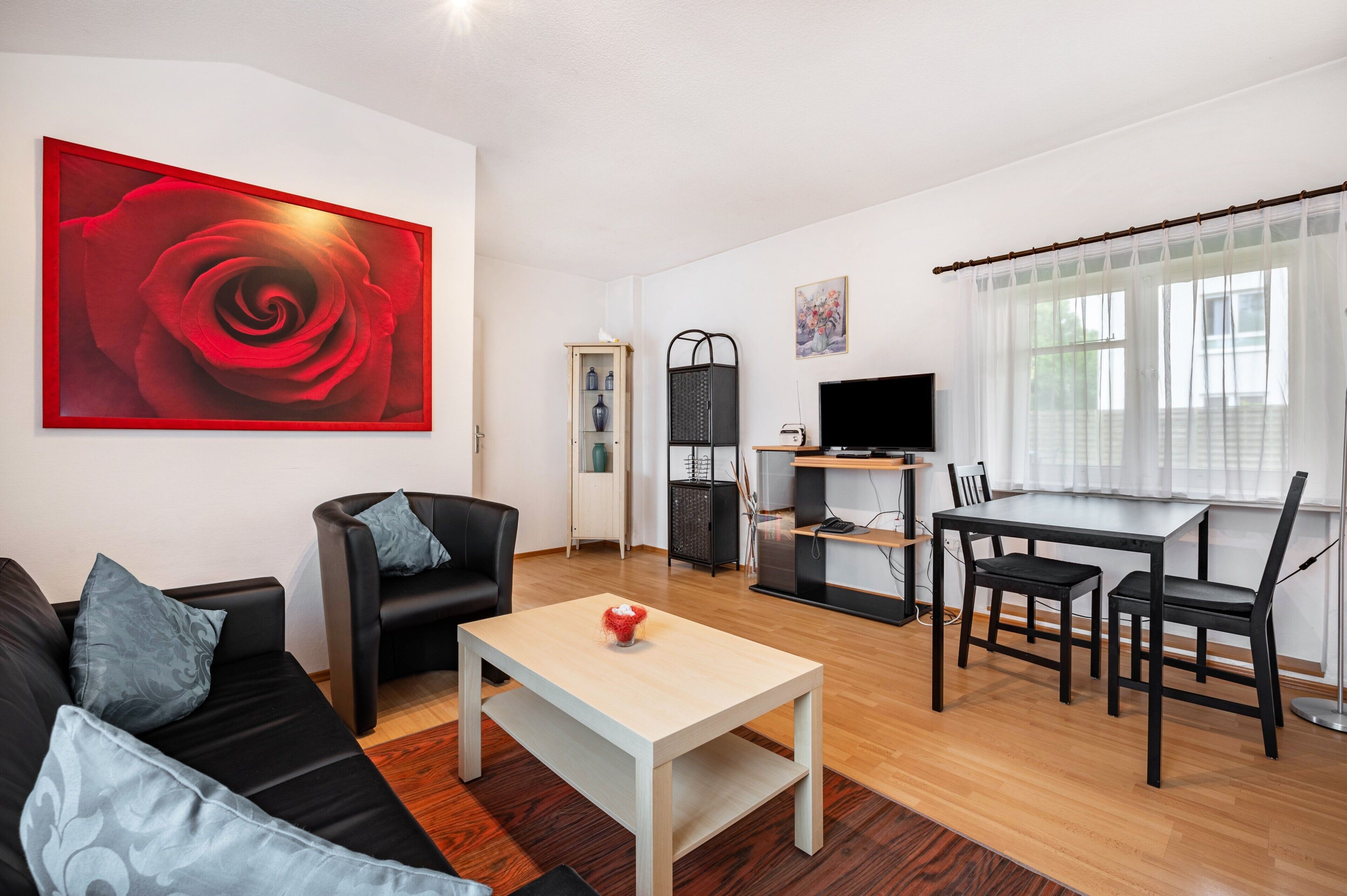 Una sala moderna con un sofá negro, una mesa de centro, una mesa de comedor con sillas, un televisor y un cuadro grande de una rosa roja en la pared.