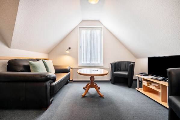 A modern loft-style room with a sloped ceiling, a black leather sofa, a round wooden table, a black armchair, a TV stand, and a window with curtains.