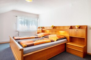 A compact bedroom with a single bed, a small bedside table, a shelf with a plant, and a window with blinds.