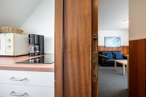Une petite cuisine équipée d’une machine à café, d’un placard et d’un comptoir. Une porte en bois donne accès à un espace de vie comprenant un canapé et une table.