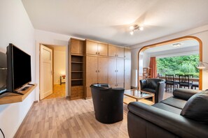 Ein modernes Wohnzimmer mit einem Flachbildfernseher, Holzkästen, einem schwarzen Mülleimer, einer Couch, einem Couchtisch und Blick auf Grünflächen durch einen Bogen.