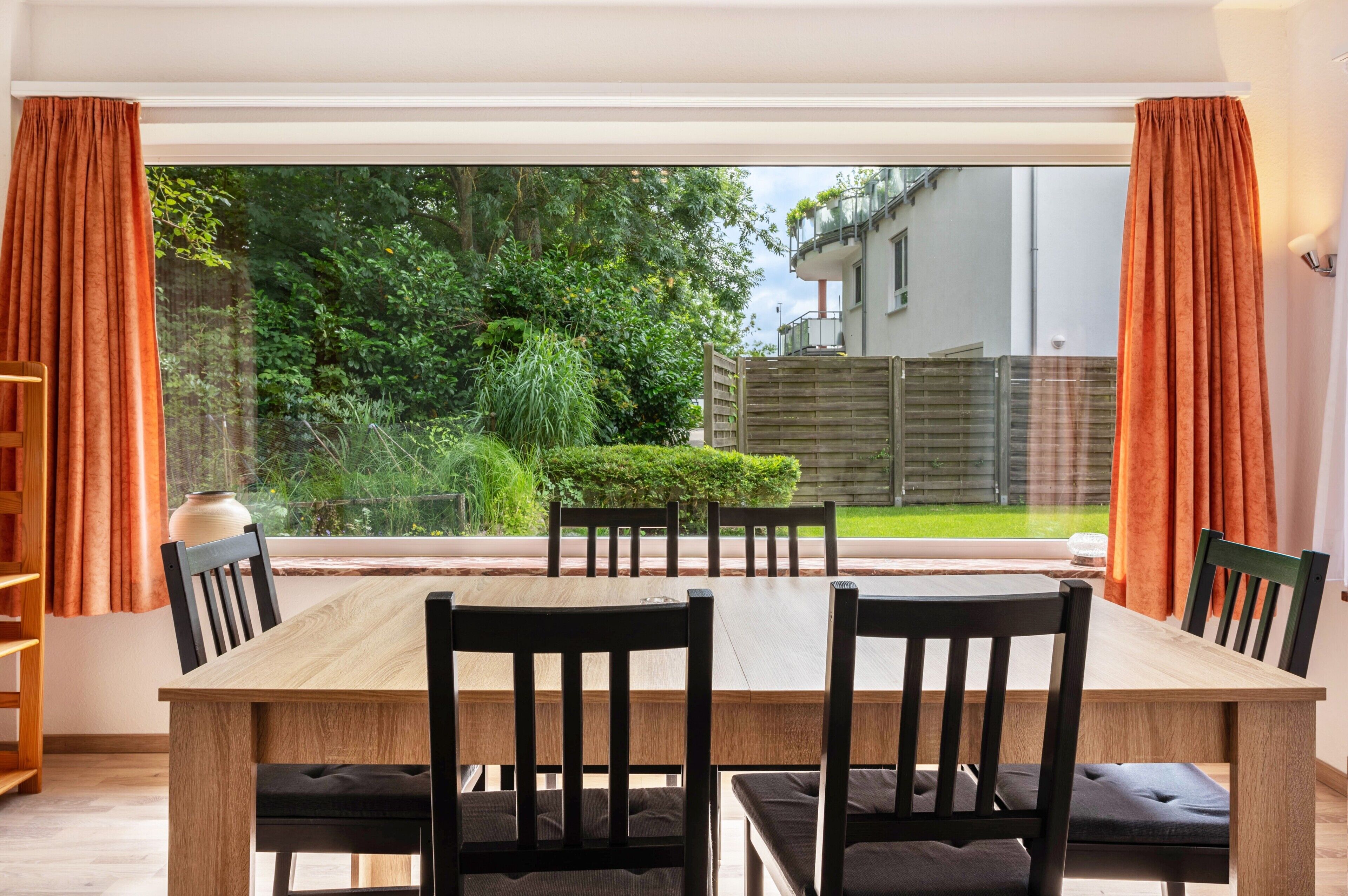 Ein Essbereich mit Holztisch und Stühlen, großen Fenstern mit Vorhängen und Blick auf Garten und Gebäude.
