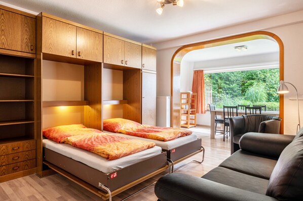 A compact room with two beds, a sofa, and a dining area with a view of greenery.