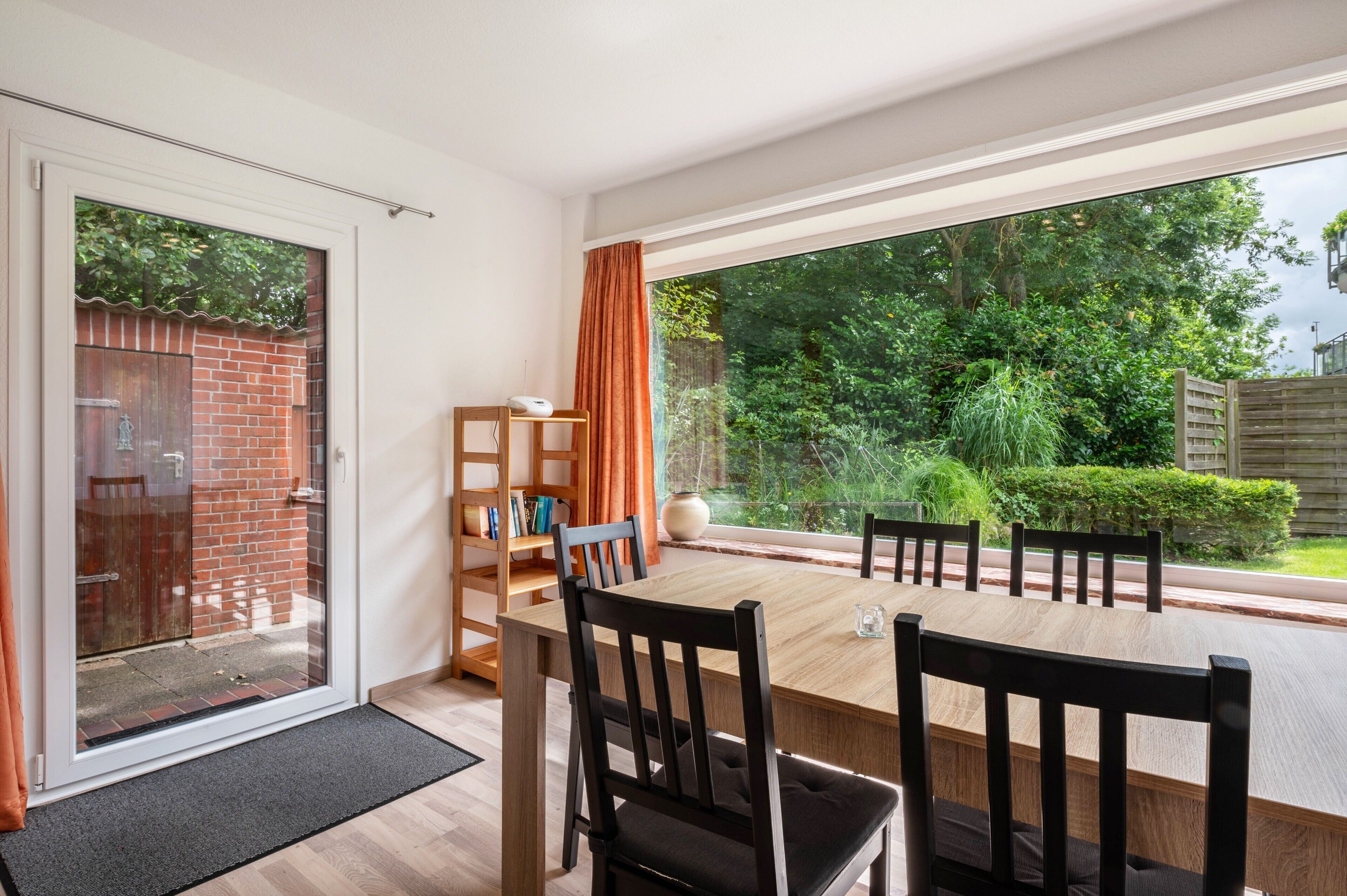 A dining area with a table, chairs, a bookshelf, and a view of a garden through a large window.