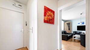 Quarto com porta branca, pintura de uma flor vermelha na parede, sofá preto, televisão e piso de madeira.