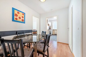 A dining area with a glass table, wooden chairs, and a blue-framed painting on the wall.