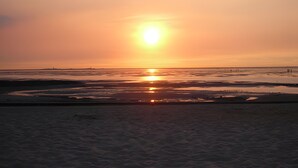 Uma praia ao pôr do sol, com o sol baixo no horizonte, águas calmas e uma linha costeira distante.