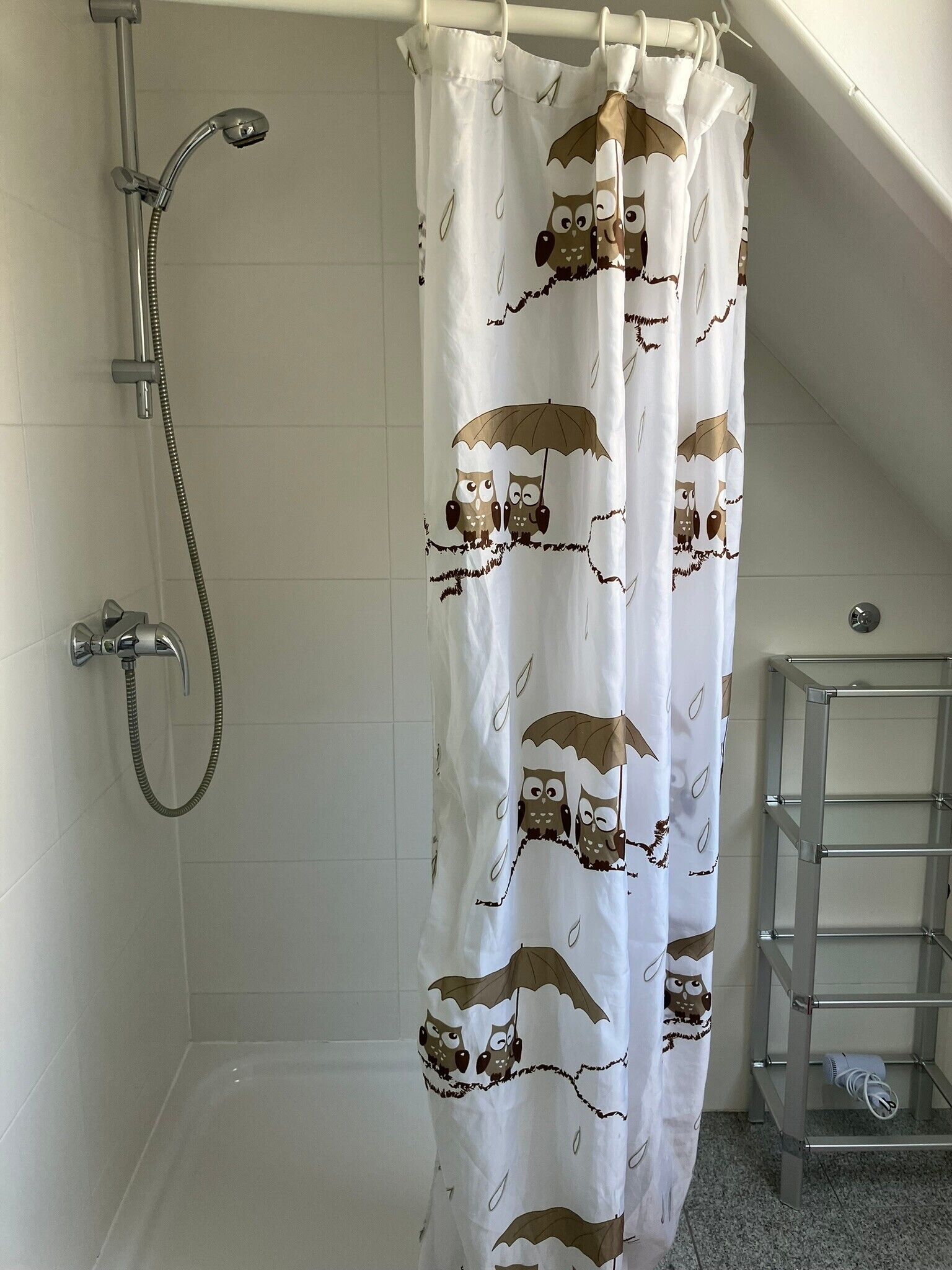 Un baño con ducha, una cortina de ducha blanca con estampado de búhos y un estante metálico.