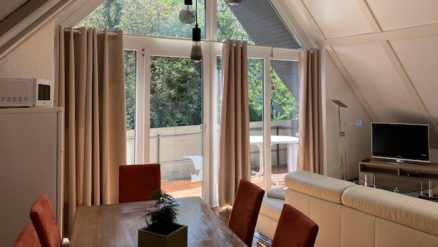 Ein Dachzimmer mit Esstisch, Stühlen, Fernseher und einem großen Fenster mit Vorhängen.