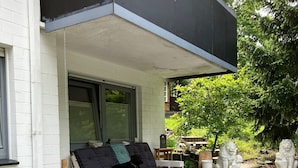 Ein überdachter Außenbereich mit einer Couch, Kissen und einem kleinen Tisch. Es befinden sich Blumentöpfe und dekorative Statuen dort.