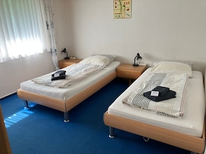 A room with two single beds, each with a nightstand, a lamp, and a small table.