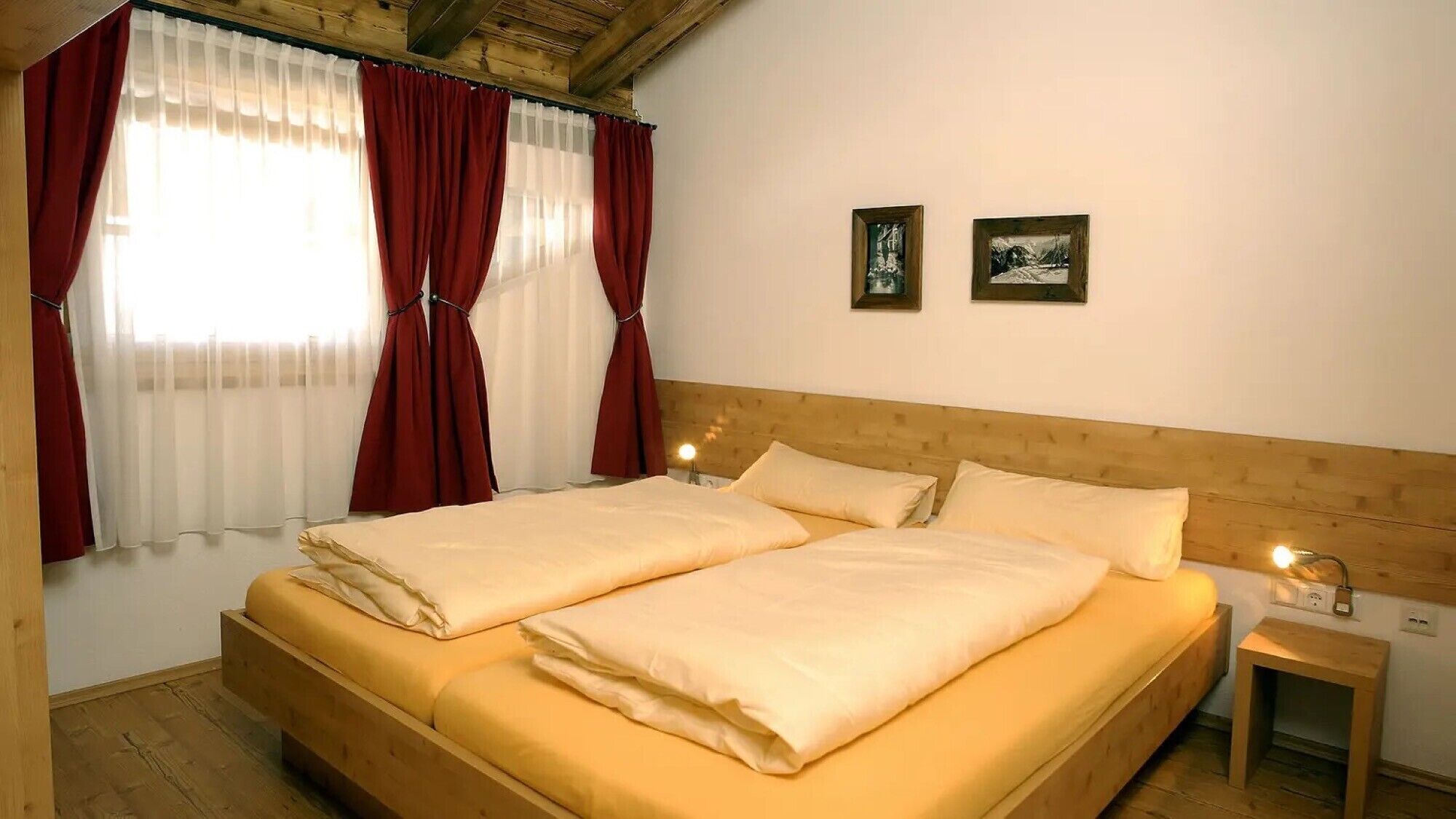 Ein Doppelbett mit Holz-Kopfteil, zwei Nachttische jeweils mit einer Lampe und ein Fenster mit roten Vorhängen.