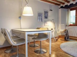 Una habitación moderna con una mesa blanca, cuatro sillas grises, una lámpara colgante, un cuadro enmarcado, una lámpara de pie y una ventana con cortinas.