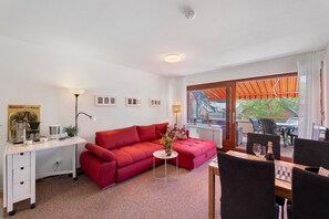 A living room with a red sofa, a small table with a vase, a desk with a lamp, and a view of the outside through a glass door.