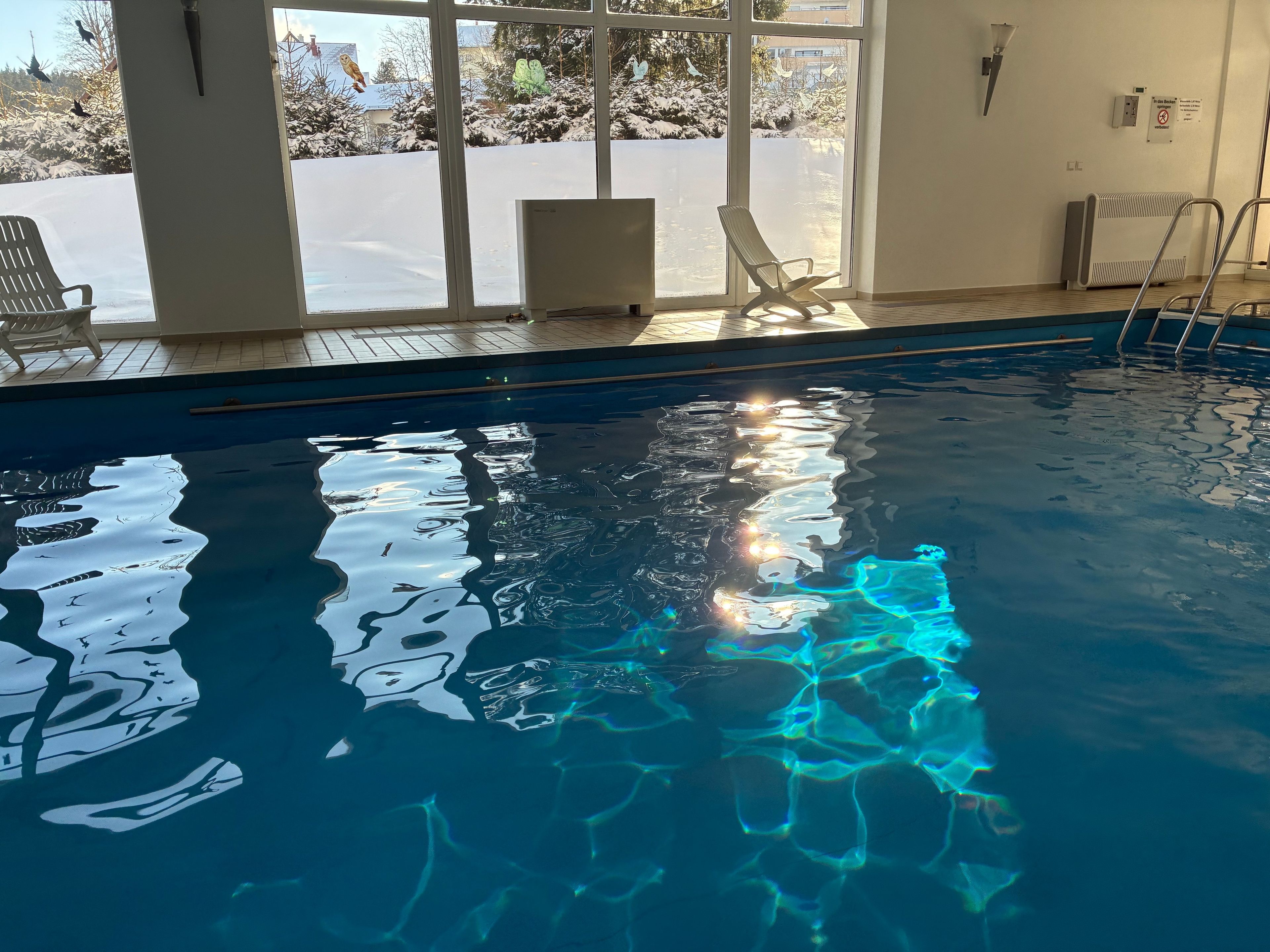 An indoor swimming pool with clear water, a view of the outside through large windows, and a chair by the poolside.
