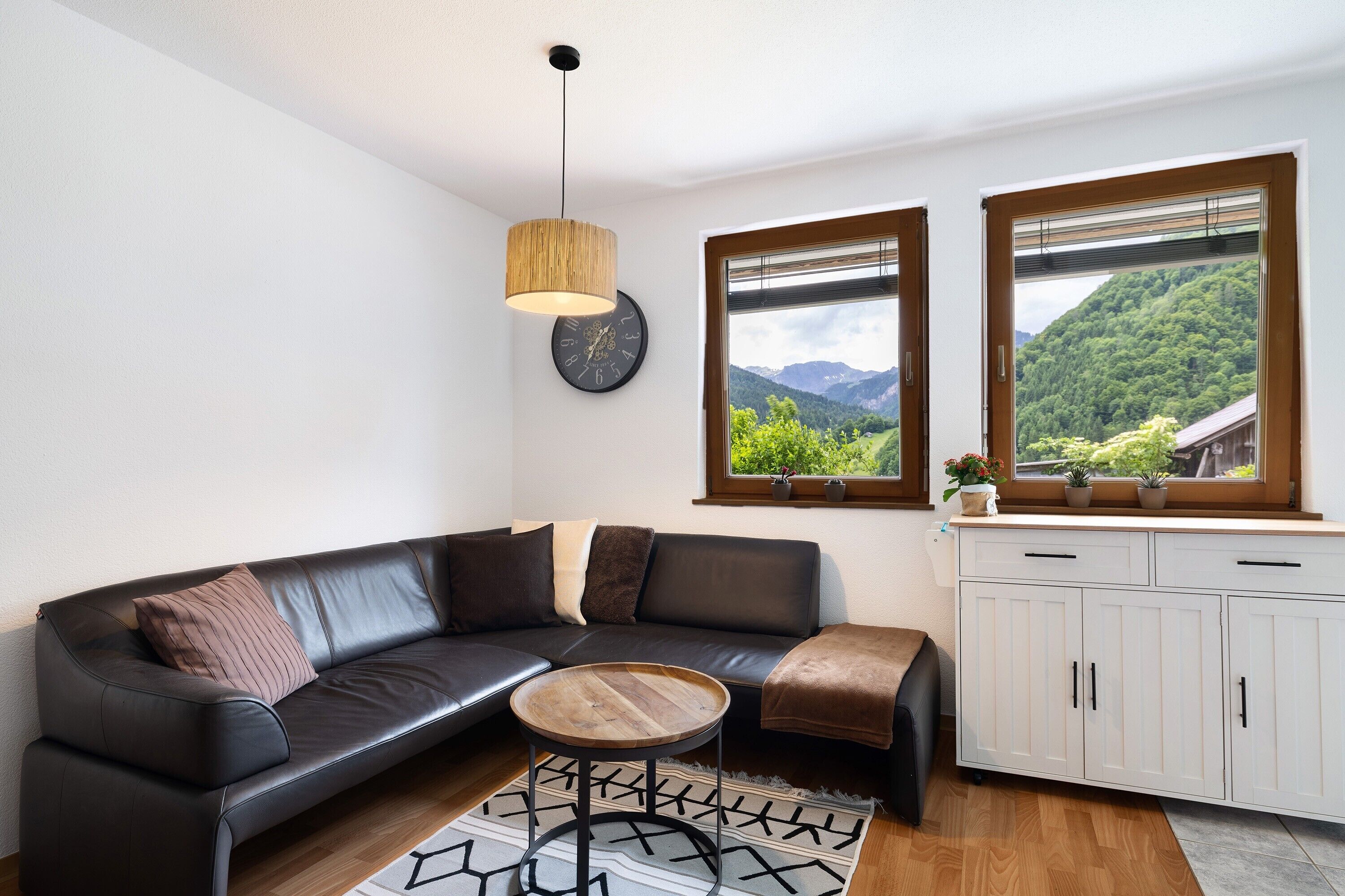 Ein modernes Wohnzimmer mit einer schwarzen Ledersofa, einem runden Holztisch, einer weißen Kommode und Blick auf eine grüne Landschaft durch große Fenster.