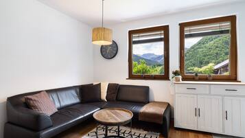 Ein modernes Wohnzimmer mit einer schwarzen Ledersofa, einem runden Holztisch, einer weißen Kommode und Blick auf eine grüne Landschaft durch große Fenster.