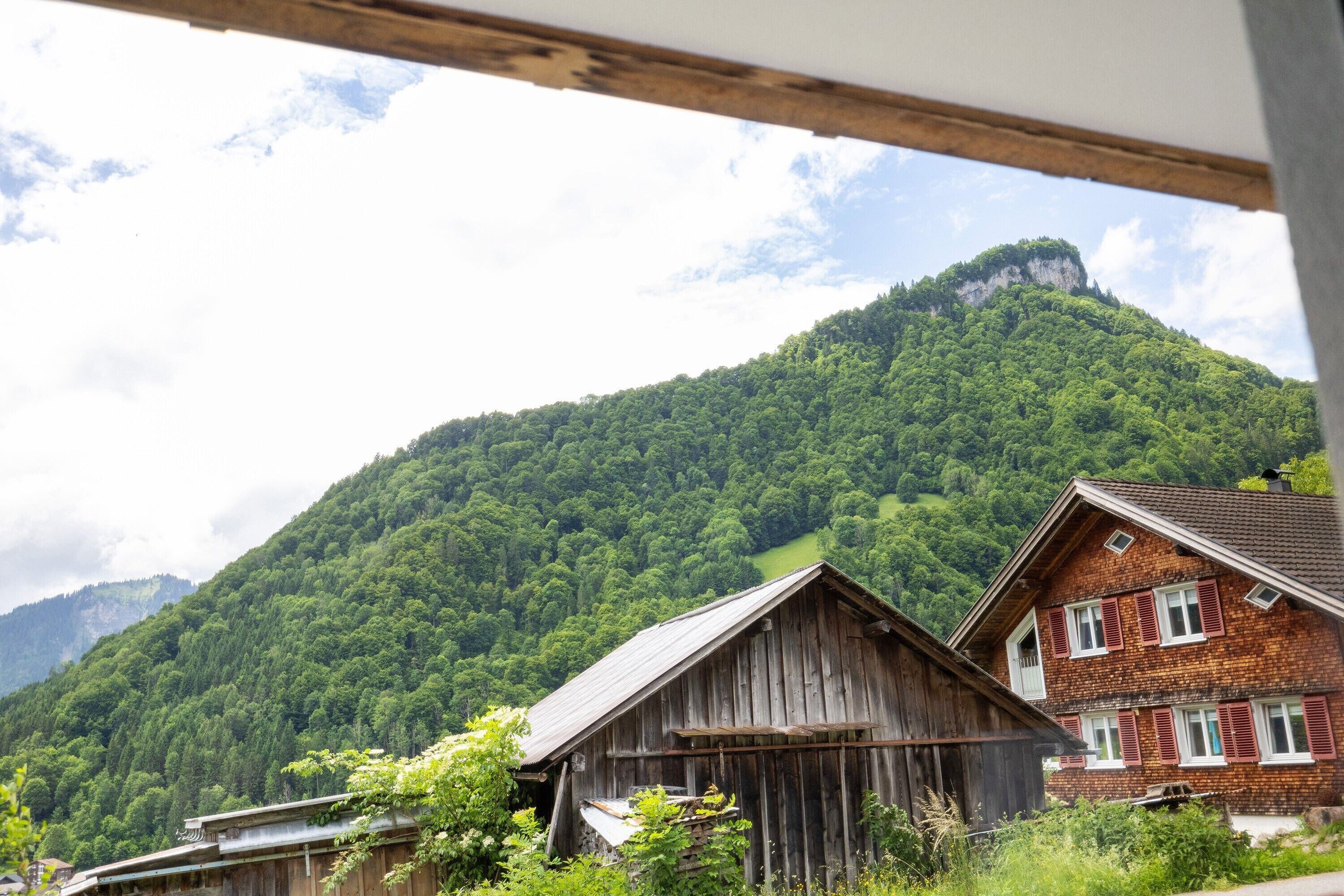A rustic wooden building with a sloped roof, surrounded by lush green hills and a mountain in the background.