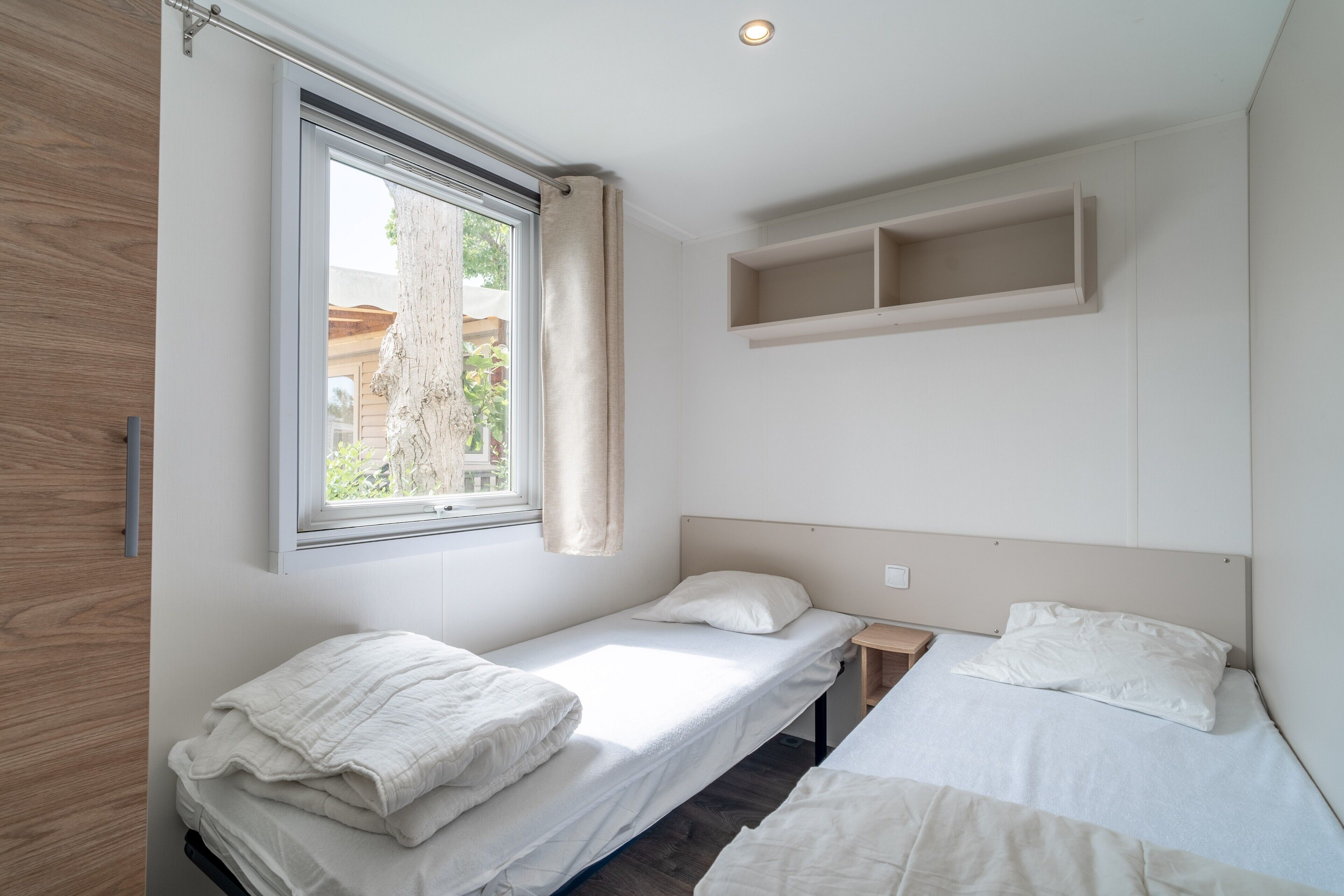 Ein Schlafzimmer mit zwei Einzelbetten, einem Fenster mit Vorhängen, einem Holzschrank und einem Regal über den Betten.