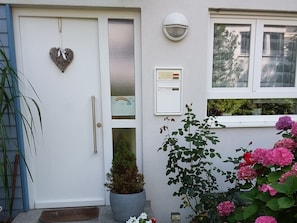 Eine weiße Tür mit Herzdeko, ein kleiner Topfpflanze und ein Fenster mit Blick auf Grünflächen.