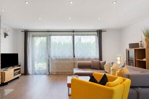Un salon moderne avec un téléviseur à écran plat, un canapé, un fauteuil jaune et une grande fenêtre avec des rideaux.