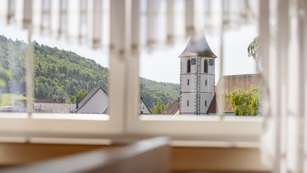 A view through a window with a wooden frame, focusing on a church with a clock tower and a forested hill in the background. - Apartment 'Waltraud' with Shared Terrace, Shared Garden and Wi-Fi (Meßstetten)