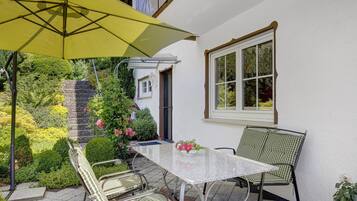 Eine Terrasse mit Tisch und Stühlen, einem gelben Sonnenschirm und einem Fenster mit Blick ins Grüne.