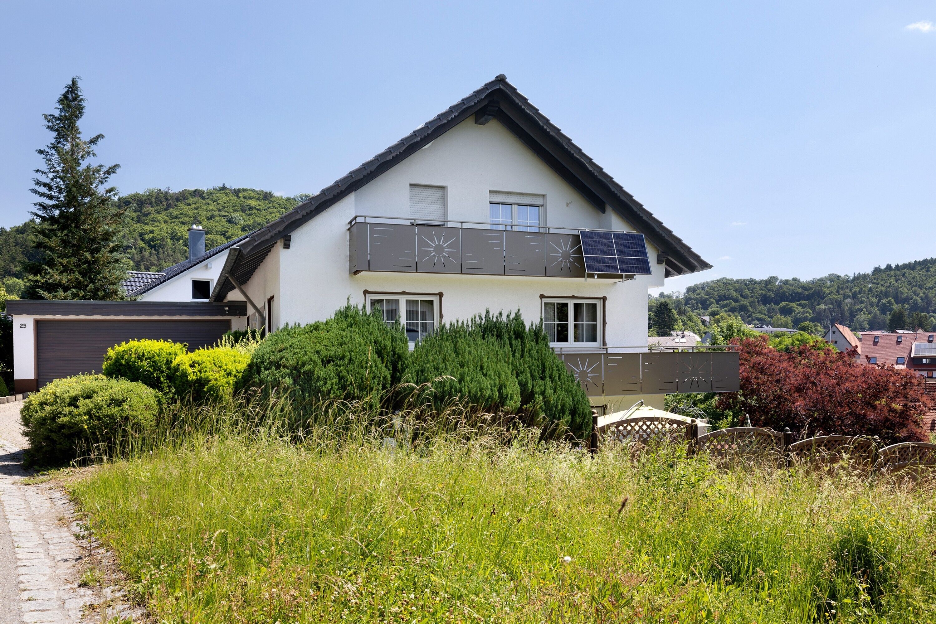 A two-story house with a balcony, surrounded by greenery and a clear sky.
