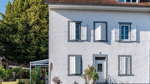 A white building with multiple windows and shutters, a small porch area, and a garden with potted plants.