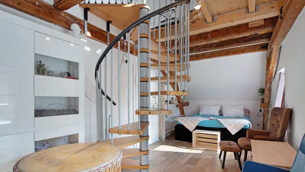 A room with a spiral staircase, a bed with a blue blanket, a wooden table, a drum, and a blue chair.