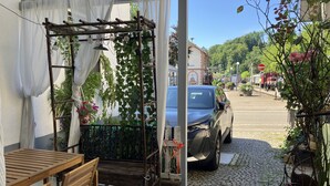 A covered outdoor seating area with a table and chairs, a wooden pergola with hanging plants, and a parked car on a cobblestone street.