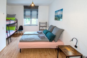 Une chambre avec des lits superposés, un bureau, une chaise, une fenêtre avec des stores et une lampe fixée au mur.