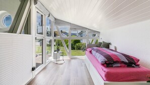 Une chambre avec une grande fenêtre, un lit recouvert d’une couverture rose et violette, et une petite table d’appoint sur laquelle est posée une plante.