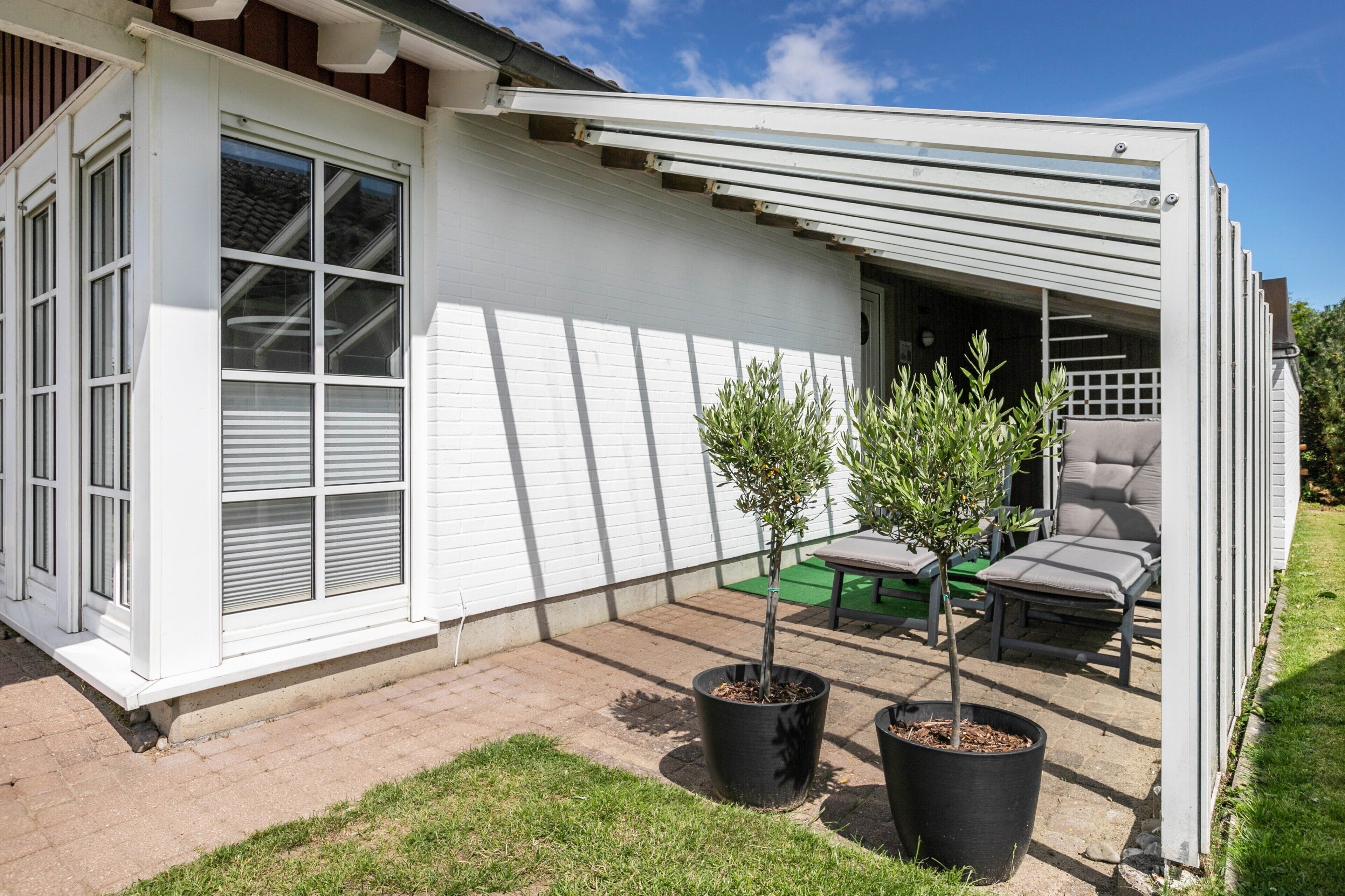 Un bâtiment blanc doté d’un patio couvert, de deux plantes en pot et de mobilier d’extérieur.
