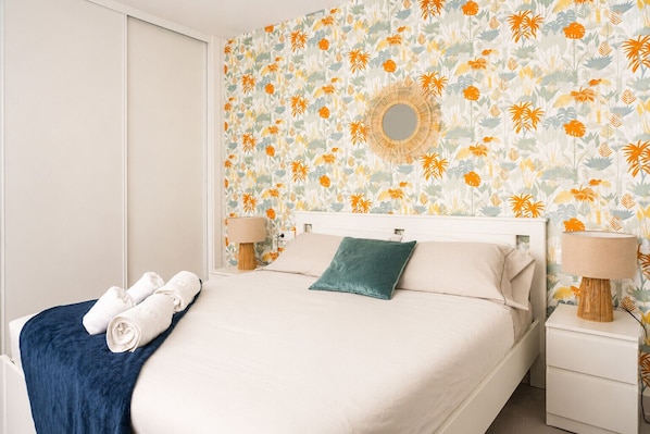 A hotel room with a bed, two bedside tables, a wall with a floral pattern, and a round mirror.