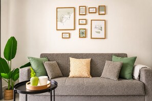 A living room with a grey sofa, green and patterned cushions, a framed artwork collection on the wall, and a plant on the side.