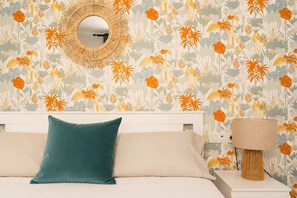 Une chambre à coucher avec un lit, un oreiller de couleur turquoise, une lampe de chevet et un mur orné d’un motif floral.
