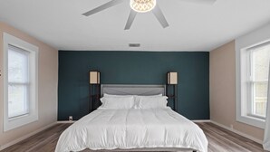 Un dormitorio con cama, dos lámparas de noche, ventilador de techo y ventana con persianas.