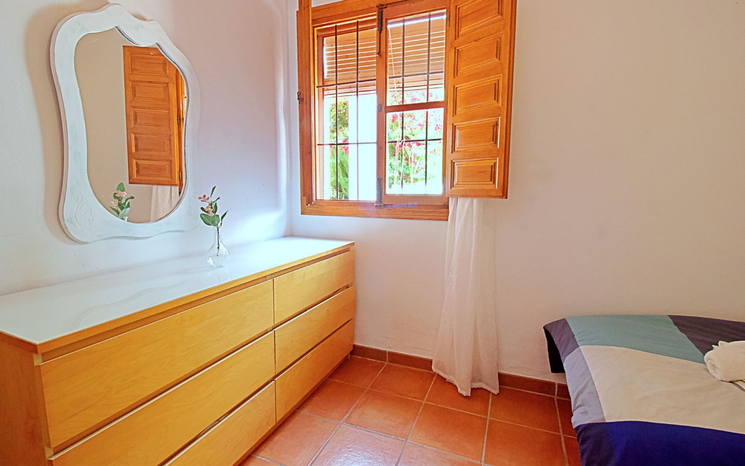 A bedroom with a wooden dresser, a bed with a quilt, a window with wooden shutters, and a mirror on the wall.