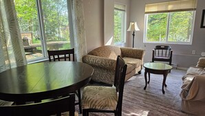 Un salon confortable avec un canapé, deux fauteuils, une petite table et une lampe. De grandes fenêtres offrent une vue sur l’extérieur.