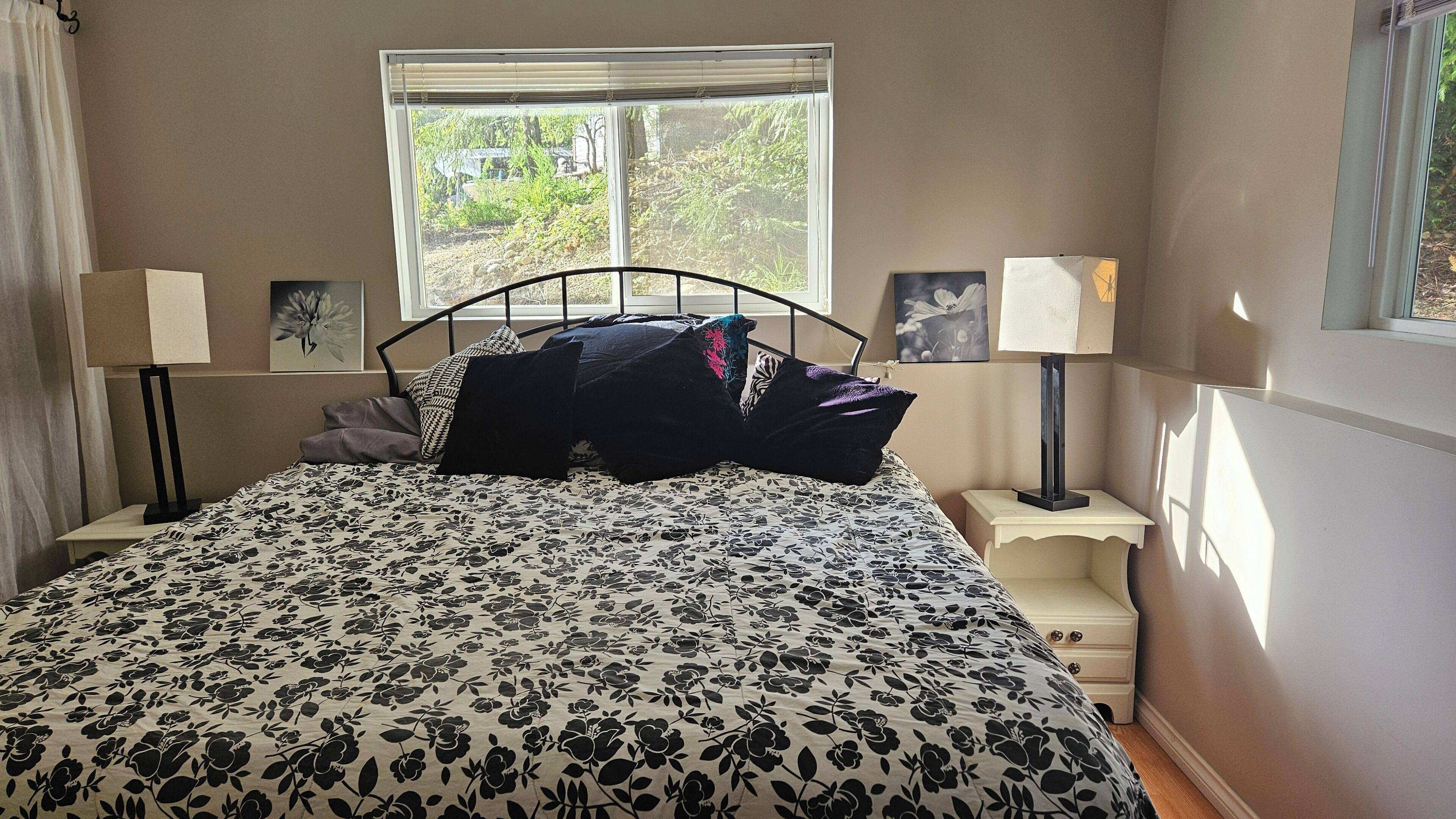 Un dormitorio con una cama, dos mesitas de noche, dos lámparas de pared, una ventana con vista a la vegetación y un cuadro en la pared.