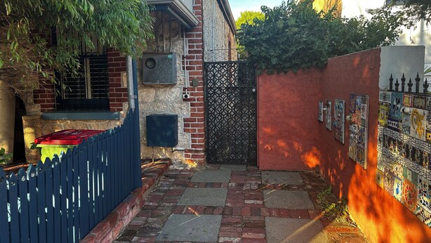 A narrow alleyway with a blue fence, a red wall with mosaic tiles, and a gate with a geometric pattern. - Stylish Studio on South terrace. 2 Minute walk to the beach. (South Fremantle)