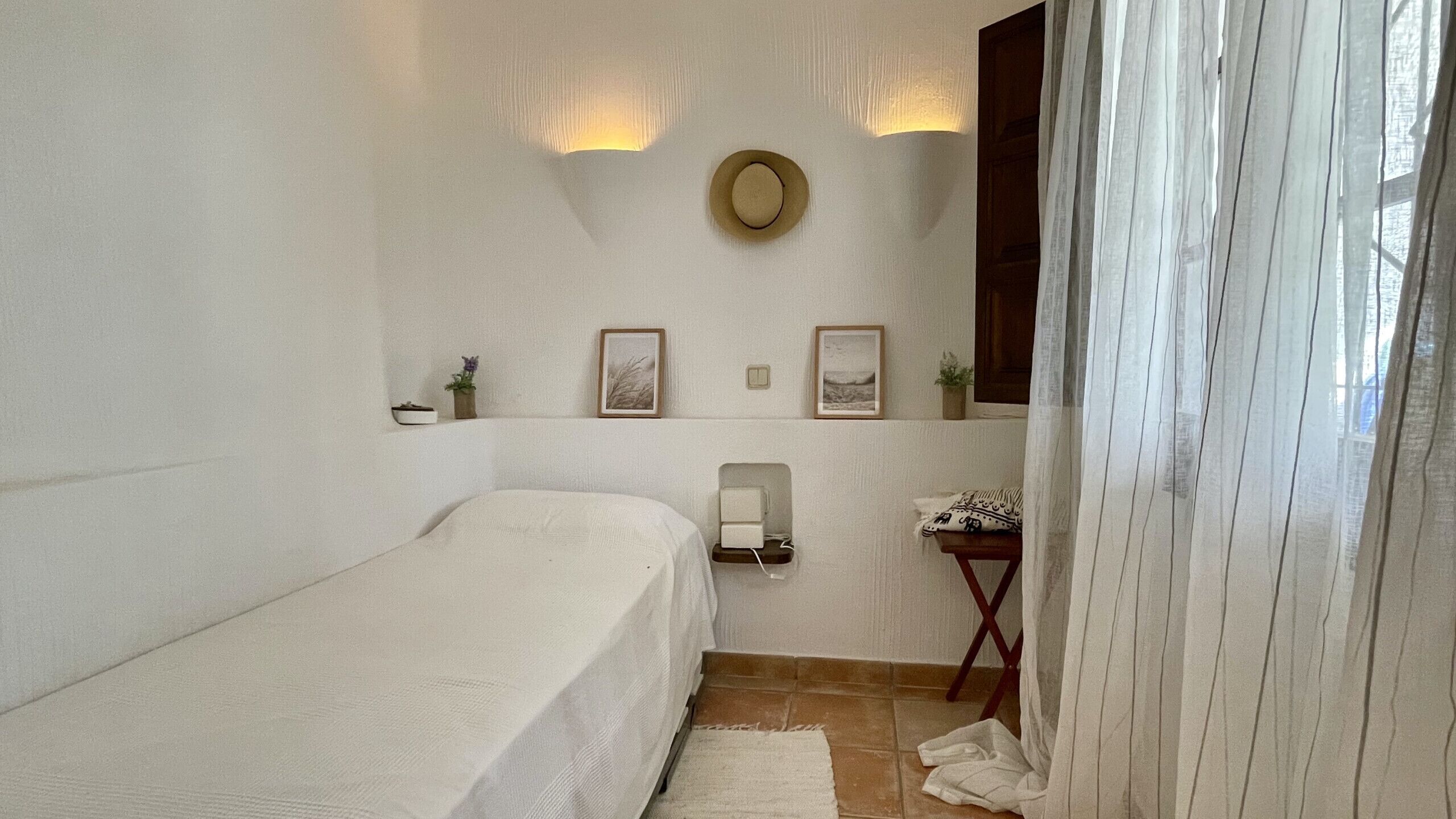 A small, neatly arranged bedroom with a single bed, a bedside table with a lamp, and a wall-mounted shelf with decorative items.