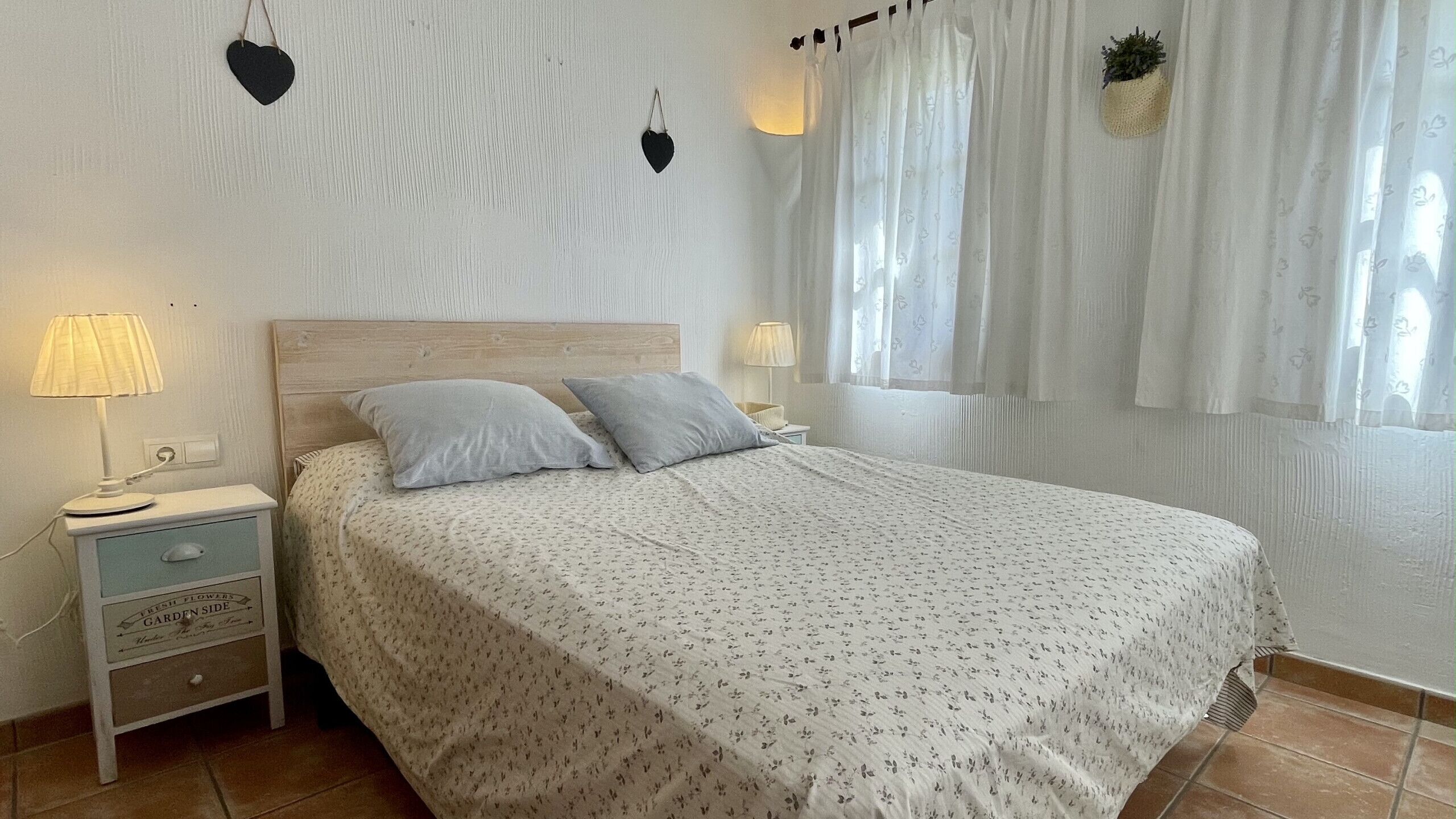 Een net opgemaakt bed met een bloemenbedekking, twee kussens, een bedlampje en een nachtkastje met een lade.