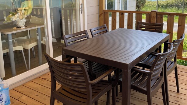 Een houten terras met een tafel en stoelen, een glazen deur naar een balkon met uitzicht op de oceaan, en een kleine tafel met stoelen binnen.